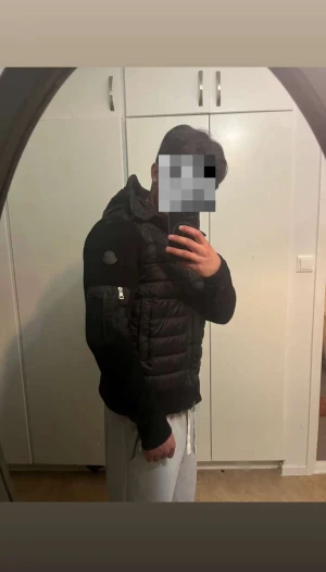 Svart Moncler pufferjacka med huva - Svart pufferjacka från Moncler med stickade ärmar och quiltad kropp. Jackan har en stor huva, dragkedja framtill och en ficka med dragkedja på ärmen. Moncler-logga på vänster ärm och snygga detaljer i metall. Perfekt för dig som vill ha en clean och modern look.