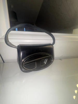 Svart handväska från Disel - Snygg svart handväska från Disel i glansigt skinn med ett stort ovalt metallspänne framtill. Väskan har en kort axelrem men det finns även en lång som kommer med och stilren design som sticker ut med sin unika detalj på framsidan. Perfekt för dig som gillar statement-accessoarer.