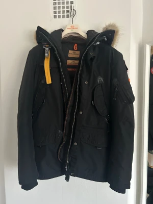 Svart Parajumpers parkajacka XL - Svart parkajacka från Parajumpers i storlek XL med päls på huvan, gul detaljrem och flera fickor med dragkedja. Jackan har slim fit-passning, quiltad insida och robusta detaljer. Perfekt för kalla dagar med snygg design och funktionella fickor.
