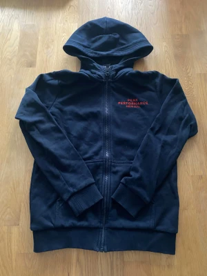 Mörkblå hoodie från Peak Performance - Mörkblå hoodie från Peak Performance med dragkedja och huva. Broderad logga i rött på bröstet. Tröjan har långa ärmar, ribbade muddar och två fickor framtill. Perfekt för chill dagar eller när du vill ha en clean och sportig look.