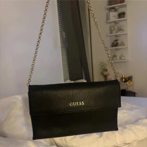 Svart axelväska från Guess - ÄKTA Snygg svart axelväska från Guess med guldfärgad kedja som axelrem och logga i guld på framsidan. Väskan har en stilren och klassisk design.💕 