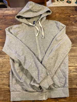 Grå hoodie med dragkedja och huva - Snygg grå hoodie med vit snörning i huvan och hel dragkedja framtill. Tröjan har långa ärmar, ribbade muddar och två fickor på sidorna. Perfekt för chill dagar och enkel att matcha med jeans eller joggers.