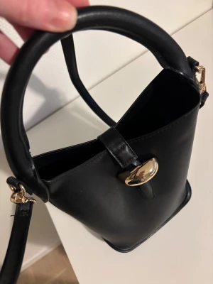 Svart bucket axelväska med gulddetaljer - Stilren svart bucket axelväska med rundad form och justerbar axelrem. Väskan har ett snyggt guldfärgat spänne framtill och detaljer i guld på remmarna. Materialet är slätt skinnliknande syntet som ger en lyxig känsla. Perfekt för dig som gillar minimalistisk design.
