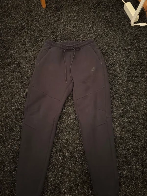 Svarta Nike tech byxor  - Svarta mjukisbyxor från Nike med dragsko i midjan och diskret Nike-logga på låret. Byxorna har en slim passform och är tillverkade i mjukt bomullsmaterial. Perfekta för chill eller träning, med praktiska fickor och snygga sömdetaljer.