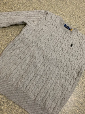 Grå stickad tröja Polo Ralph Lauren - Snygg grå kabelstickad tröja från Polo Ralph Lauren med klassisk rund hals och broderad logga på bröstet. Tröjan har långa ärmar och ribbade muddar vid ärmslut och nederkant. Perfekt för dig som gillar stilren och tidlös design. Taggar finns fortfarande på, glömde ta kort på dom 