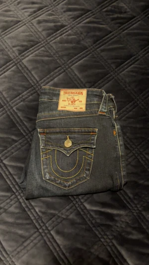 True Religion mörkblå bootcut jeans - Mörkblå jeans från True Religion med klassiska kontrastsömmar och stora fickor med lock och knapp bak. Bootcut-modell med normal midja och snygg passform. Ikonisk logga på bakfickan och patch i midjan. De är i jättebra skick.💖