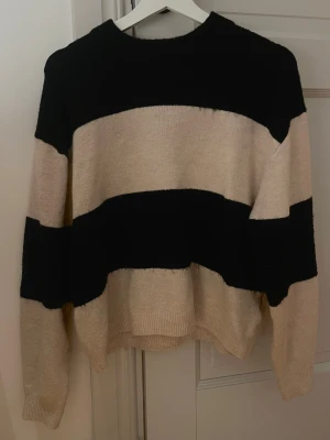 Randig stickad tröja i svart och beige - Säljer en chill stickad tröja med breda svarta och beige ränder. Tröjan har en relaxed passform, rund hals och långa ärmar. Perfekt för att hålla sig varm och se snygg ut under hösten och vintern.