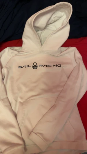 Vit hoodie från Sail Racing - Vit hoodie från Sail Racing med svart logga tryckt på bröstet och på huvan. Klassisk känguruficka framtill och mjuk insida. Perfekt för dig som gillar stilren och sportig look. Snygg passform och skön att bära.