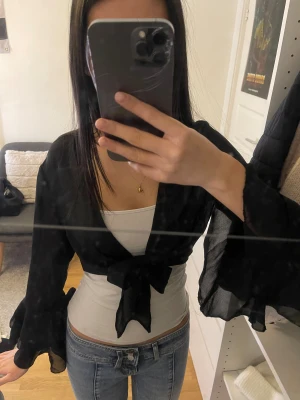 Svart bolerotopp med knyt - Säljer en svart bolerotopp i tunn, luftig polyester med stora volangärmar och knyt framtill. Toppen är croppad och har en snygg öppen design som passar över linnen eller klänningar. 💕Skriv för frågor, kolla min profil för annat snyggt