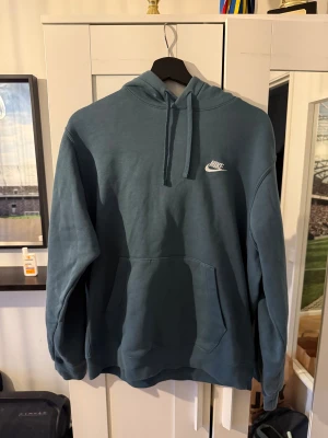Blå Nike hoodie med logga - Säljer en blå hoodie från Nike med vit broderad logga på bröstet. Tröjan har huva med snörning och en stor magficka framtill. Perfekt för chill dagar och enkel att matcha med jeans eller joggers.
