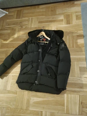 Svart pufferjacka från Moose Knuckles - Mörkblå/svart pufferjacka från Moose Knuckles med huva, silverfärgade knappar och dragkedja. Jackan har flera fickor med lock och en snygg logga på ärmen. Insidan är mörkblå och fodrad, perfekt för kalla dagar. Stilsäker och rejäl design. Säljs pågrund av att har köpt nytt vinterjacka har använts 2-3 gånger Storlek L/XL