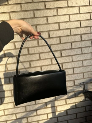 Svart axelväska - Stilren svart axelväska i med minimalistisk design och rektangulär form. Väskan har ett smalt axelband och passar perfekt för dig som gillar en clean och klassisk look. Perfekt storlek för det viktigaste och enkel att matcha med olika outfits. Använd ett fåtal gånger! 