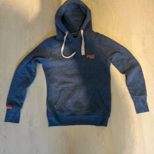 Blå hoodie från Superdry - Säljer en blåmelerad hoodie från Superdry Orange Label med vit snörning i huvan och broderad logga på bröstet. Tröjan har känguruficka framtill och ribbade muddar. Perfekt för chill dagar och snygg streetstyle.
