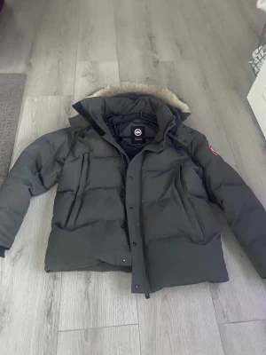 Mörkgrön Canada Goose pufferjacka XL - Säljer en mörkgrön pufferjacka från Canada Goose i storlek XL. Jackan har en stor huva med päls, klassisk logga på ärmen och dragkedja med knappar framtill. Perfekt för kalla vinterdagar och har en riktigt schysst streetstil.