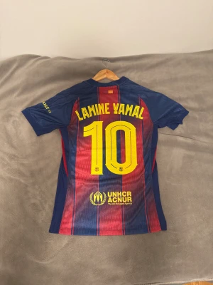 Lamine Yamal fotbollströja  - Säljer en officiell FC Barcelona fotbollströja med Lamine Yamal och nummer 10 på ryggen. Tröjan har klassiska blå och röda vertikala ränder, gula tryck och UNHCR- loga. PRIS KAN DISKUTERAS