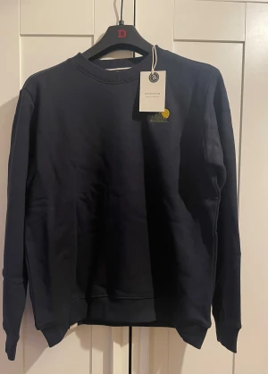 Mörkblå sweatshirt från Anerkjendt - Snygg mörkblå sweatshirt från Anerkjendt med broderad detalj av berg och sol på bröstet. Tröjan har rund hals, ribbade muddar och är tillverkad i mjuk bomull. Perfekt för dig som gillar stilrena och coola plagg med en twist.