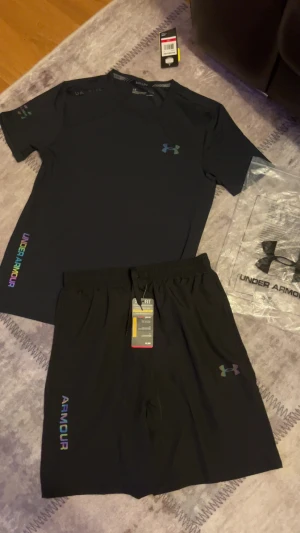 Svart Under Armour t-shirt, Shorts - Svart Under Armour t-shirt med korta ärmar och färgglada logodetaljer på bröstet och sidan. Materialet är lätt och andas, perfekt för träning. Snygg, sportig design med UA-logga i regnbågsfärger och rund hals. Passar dig som gillar stilren och funktionell sportklädsel.