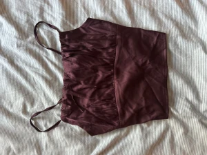 Vinröd croppad linnetopp med justerbara band - Säljer en vinröd croppad linnetopp med smala axelband och snygga rynkade detaljer framtill. Toppen har ett knytband i ryggen som ger en extra cool touch. Materialet är glansigt och känns mjukt mot huden, perfekt för att lyfta din outfit.