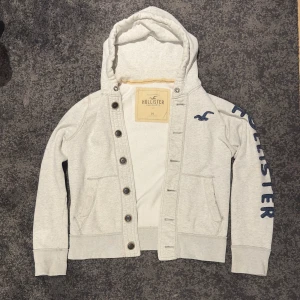 Ljusgrå Hollister Cardigan - Säljer denna eftertraktade Hollister cardigan. Den är grå med blåa detaljer. Storlek M och bra skick. Finns en liten flack vid S vid texten men det går säkert att tvätta bort. OBS: Tvättas inte innan frakt.
