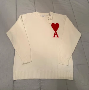 Vit stickad tröja från Ami Paris - Vit stickad tröja från Ami Paris med röd hjärt- och A-logga på bröstet. Rund hals och ribbade muddar vid ärmslut och nederkant. Tröjan har lång ärm och är tillverkad i mjuk ull, perfekt för att hålla dig varm och snygg under kyliga dagar.