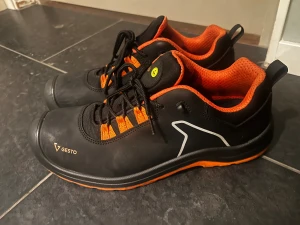Svarta och orange arbetsskor från Gesto - Säljer ett par svarta sneakers från Gesto med coola orange detaljer och meshfoder. Skorna har snörning, slitstark platt sula med orange undersida och rund tå. Perfekt för dig som gillar sportig stil och vill sticka ut med färg. Märkning och storlek syns tydligt på plösen.