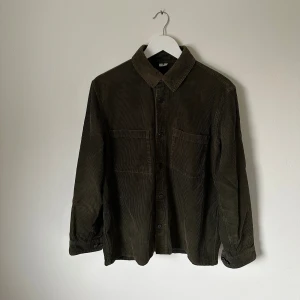 Olivgrön manchester överskjorta från Arket - Olivgrön överskjorta i manchester från Arket med klassisk krage, två bröstfickor och knäppning framtill. Skjortan är långärmad och har en loose fit, perfekt för lager-på-lager. Tillverkad i bomullscorduroy som ger en snygg retrovibe. 5G7