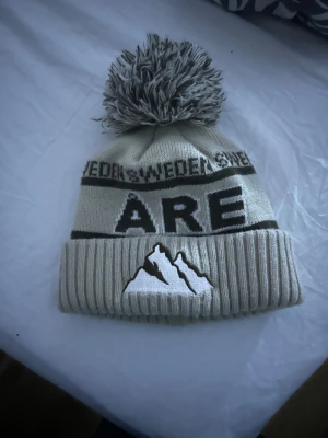 Beige stickad Åre mössa med tofs - Säljer en beige stickad mössa med stor tofs på toppen och ribbad kant. Mössan har texten 'SWEDEN' och 'ÅRE' samt ett broderat bergmotiv framtill. Perfekt för kalla dagar och vinterhäng i fjällen.