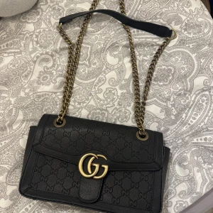 Svart Gucci axelväska med kedja - Snygg svart axelväska från Gucci med GG-logga i guld framtill och präglat GG-mönster över hela väskan. Väskan har en kraftig guldfärgad kedja som axelrem och insida i beige. Praktisk dragkedja på insidan för småsaker.