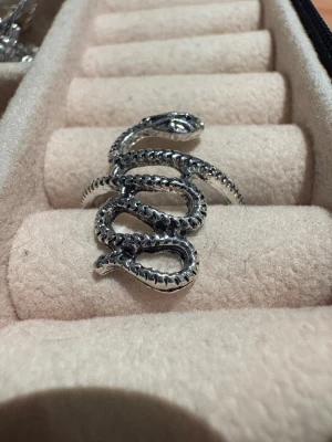 Silvrig ring med orm-design - Cool ring i silverfärgad metall formad som en slingrande orm. Detaljerad yta med markerade fjäll och ett upphöjt ormhuvud som ger en edgy vibe. Perfekt för dig som gillar unika och djur-inspirerade smycken.