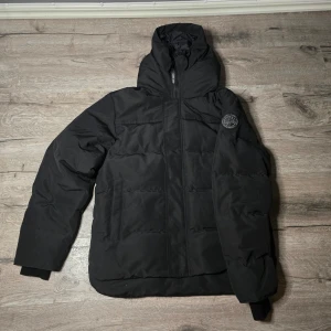  Canada Goose jacka - Svart pufferjacka från Canada Goose med huva och diskret logotyp på ärmen. Jackan har dragkedja framtill och quiltad design som ger extra värme. Medföljer originalpåse och tags. Perfekt för kalla dagar och har en snygg, modern look.jackan e helt ny. Den var tyvärr för stor för mig! Akopia 