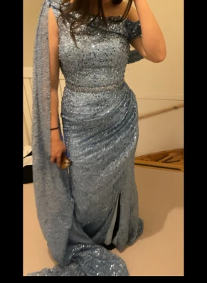 Blå glittrig långklänning med släp och uppet vid högra sidan  - Magisk blå långklänning med glittriga paljetter över hela tyget, elegant släp och en axelbandslös design med draperad effekt. Klänningen har markerad midja med glittrigt bälte och en snygg slits framtill. Perfekt för bal eller festkvällar när du vill sticka ut.