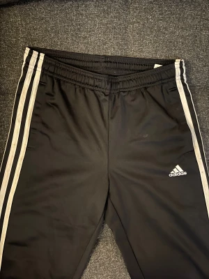 Svarta Adidas träningsbyxor  - Svarta träningsbyxor från Adidas med vita ränder längs sidorna och Adidas-logga på benet. Byxorna har elastisk midja.