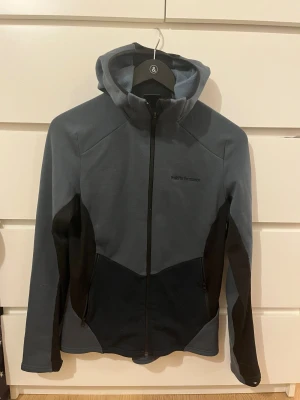 Peak Performance blå fleecejacka - Peak Performance hoodiejacka i mörkblå och svart färg, med hel dragkedja och två sidofickor. Jackan har mjukt fleecefoder på insidan och broderad logga på bröstet. Snygg passform och perfekt för kyliga dagar. Långärmad och med huva för extra komfort. I storlek M för barn
