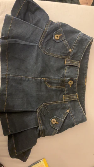 Mörkblå jeanskjol med fickdetaljer - Snygg mörkblå jeanskjol med markerade sömmar och coola fickdetaljer med knappar framtill. Kjolen har en lätt utställd form och kort längd, perfekt för en trendig look. Klassisk denim med gult kontraststick, passar till många stilar.