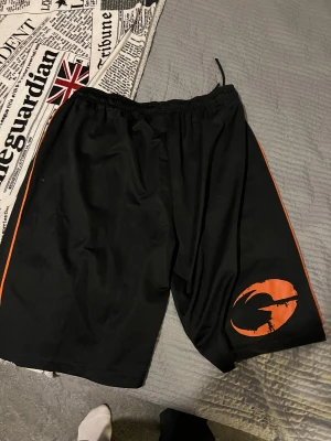 GASP svarta shorts - Aldrig använda svarta träningsshorts från GASP med orangea detaljer längs sidorna och stort tryck på ena benet. Shortsen har snörning i midjan och är gjorda i ett lätt och ventilerande syntetmaterial, perfekt för gymmet eller sport. Snygg och sportig look med logga framtill.