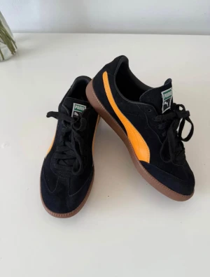 Svarta Puma sneakers med orange detalj - Säljer ett par snygga Puma sneakers i svart mocka med klassisk orange sidostrip och brun gummisula. Skorna har snörning och rund tå, samt Puma-logga på plösen och yttersulan.    «På sidan finns det ingen möjlighet att välja min faktiska storlek, därför anger jag i beskrivningen: skostorlek 41 EU, fotlängd 26,5 cm.» ❗️