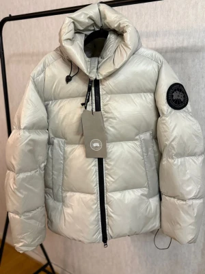 Ljusgrå  Canada Goose - Snygg ljusgrå pufferjacka från Canada Goose med stor huva, svarta detaljer och patch på ärmen. Jackan har dragkedja framtill, två stora fickor och justerbar snörning vid huvan. Insidan har svarta axelband med logga och meshfickor. Helt ny finns I flera storlekar.