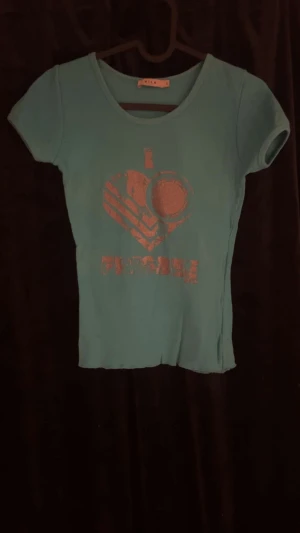 Turkos ribbad topp från Vila - Turkos ribbad topp från Vila i storlek S. Toppen har korta ärmar och ett tryck framtill med texten 'I love frisbee' i silver. Rund halsringning och figurnära passform. Perfekt för dig som gillar färgglada plagg.