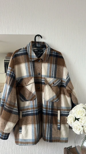 Rutig overshirt jacka från Zara - Snygg rutig overshirt jacka från Zara med stora bröstfickor och knappar framtill. Färgerna är beige, brun, blå och vit, vilket ger en chill vibe. Jackan är gjord i ullblandning och har en loose passform med klassisk krage.