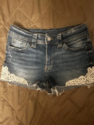Blå jeansshorts med spetsdetaljer - Snygga blå jeansshorts med fransig kant och vita spetsdetaljer, Passar inte längre så provar lägga upp dom här💕