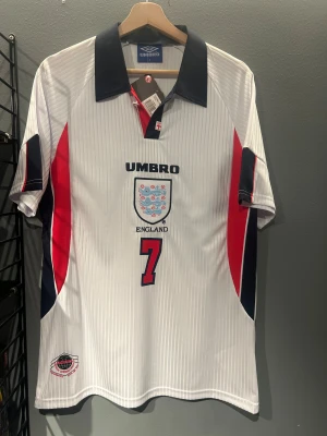 England Beckham #7 fotbollströja Umbro - Säljer en klassisk England fotbollströja från Umbro med Beckham och nummer 7 på ryggen. Tröjan är vit med tunna vertikala ränder, har mörkblå krage och detaljer i rött och mörkblått på sidorna och ärmarna. Broderat England-emblem på bröstet. Materialet är polyester. Helt ny tröja en replika