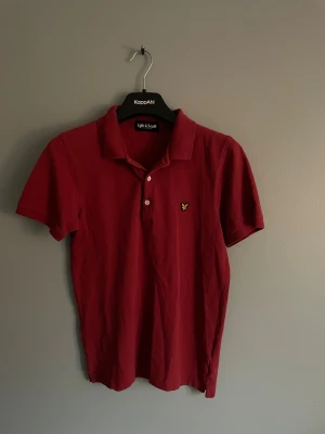Röd pikétröja från Lyle & Scott - Röd pikétröja från Lyle & Scott med klassisk krage och tre vita knappar framtill. Tröjan har korta ärmar och den ikoniska gula örnloggan broderad på bröstet. Tillverkad i mjuk bomull som är skön mot huden.