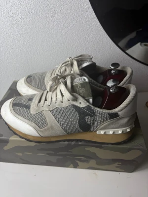Valentino Rockrunner sneakers grå/vita - Säljer ett par Valentino Rockrunner sneakers i grått, vitt  och beige med coolt kamouflagemönster. Skorna har snörning, platt sula och detaljer i mocka och mesh. Ikoniska nitar vid hälen och svart yttersula. Kommer med originalkartong, extra skosnören och dustbag.