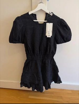 Svart kort klänning från Zara XS - Supersöt svart klänning från Zara i storlek XS. Klänningen har puffiga korta ärmar, broderade detaljer och en volangkant nedtill. Smockad midja ger en snygg siluett och den har rund hals. Perfekt för dig som gillar stilrena och trendiga plagg. Helt ny med prislapp!!!