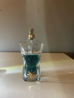 Jean Paul Gaultier Le Male parfym - Ikonisk parfymflaska formad som en manlig torso från Jean Paul Gaultier. Flaskan är gjord av genomskinligt glas med blågrön vätska inuti och har guldfärgad spraykork. Dekorativ detalj i guld vid basen. Perfekt för dig som gillar unika designer.