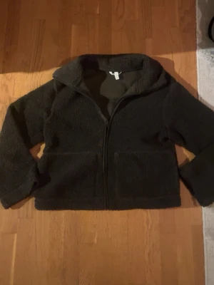 Mörkgrön teddyjacka med dragkedja - Mysig mörkgrön teddyjacka med dragkedja framtill och två stora fickor. Jackan har en hög krage och är långärmad med en loose passform. Perfekt för att hålla värmen under kyliga dagar.