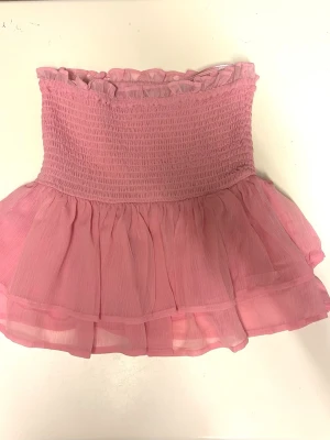 Rosa volang tubtopp - Supersöt rosa tubtopp med smockad överdel och volangig nederdel. Toppen är ärmlös och har en romantisk känsla med volanger upptill och nertill. Perfekt för dig som vill ha en feminin och trendig look.