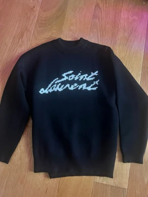 Svart stickad Saint Laurent-tröja - Snygg svart stickad tröja från Saint Laurent med vit logotyp i grafisk stil på bröstet. Tröjan har rund halsringning och ribbade muddar vid ärmslut och nederkant. Perfekt för dig som gillar  trendiga plagg..Storlek S,loose passform,passar båda S/M