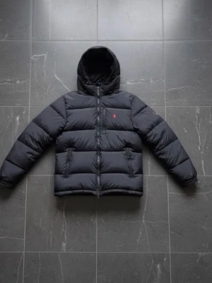 Svart dunjacka från Polo Ralph Lauren - Snygg svart dunjacka från Polo Ralph Lauren med huva och diskret röd logga på bröstet. Jackan har quiltad design, dragkedja framtill och två sidofickor. Perfekt för kalla dagar och ger en clean, modern look.