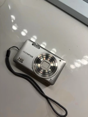 Olympus silverfärgad digitalkamera - Snygg och kompakt digitalkamera från Olympus i silverfärgad metall. Kameran har en stor display på baksidan, 5x optisk zoom och HD Movie-funktion. Utrustad med smidigt handledsband och flera knappar för enkel navigering. Perfekt för att fånga minnen med stil. Om man vill så betalar man extra 100kr för SD Kort 